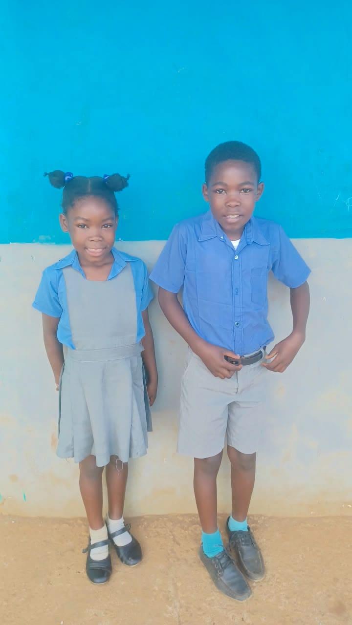Two children in school uniform, a girl in a grey pinafore and a boy in a blue shirt and grey shorts, standing together against a painted wall.